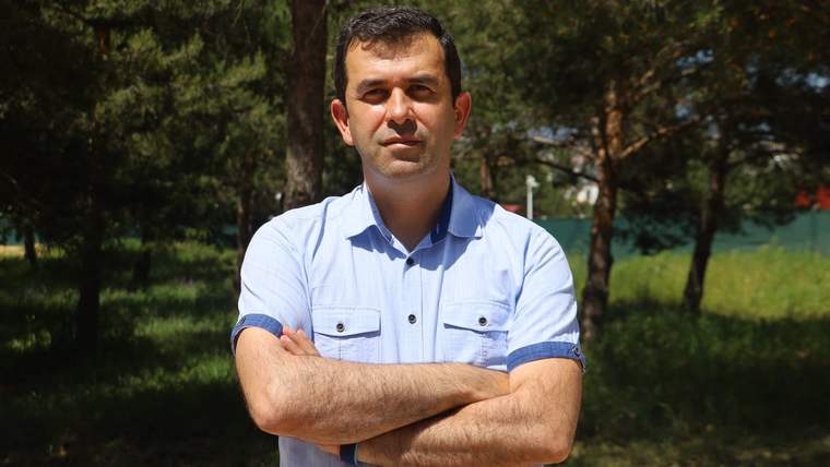 Ağaçlık bir alanda, kollarını göğsünde kavuşturmuş kısa saçlı bir kişi duruyor.