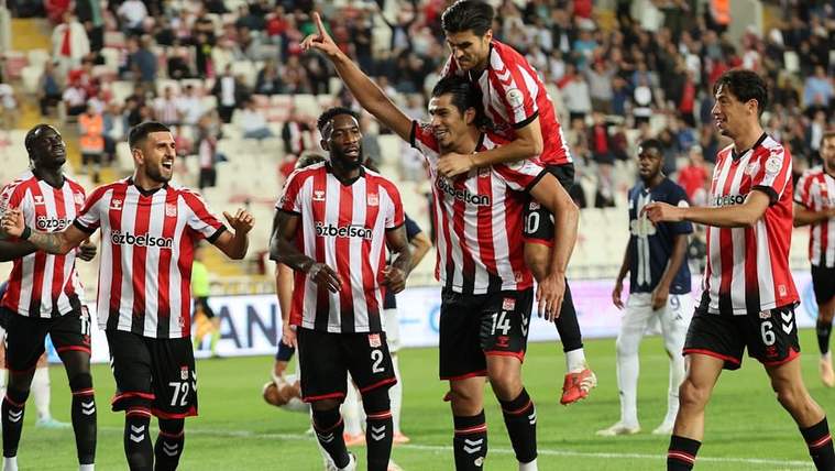 Kırmızı beyaz formalar giyen futbolcular sahada sevinirken bir oyuncu takım arkadaşını sırtında taşıyor.