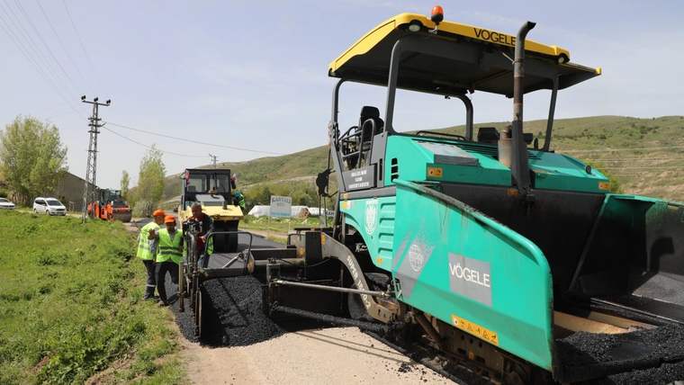 Kırsal yolda sıcak asfalt seren Vögele marka asfalt finişeri ve yanında çalışan işçiler görülüyor.
