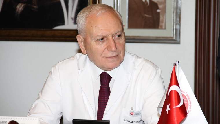 Beyaz önlüklü bir kişi masa başında oturuyor; yanında Türk bayrağı ve beyaz renkli bir flama bulunuyor.