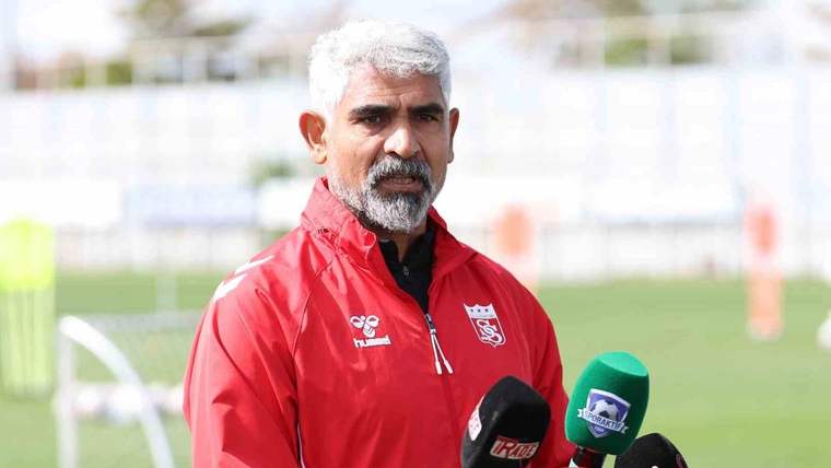 Yeşil saha önünde kırmızı spor ceketli bir kişi, mikrofonların karşısında konuşuyor.