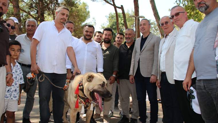 Ağaçların arasında bir araya gelen kalabalık grup, tasmalı büyük bir köpekle birlikte poz veriyor.