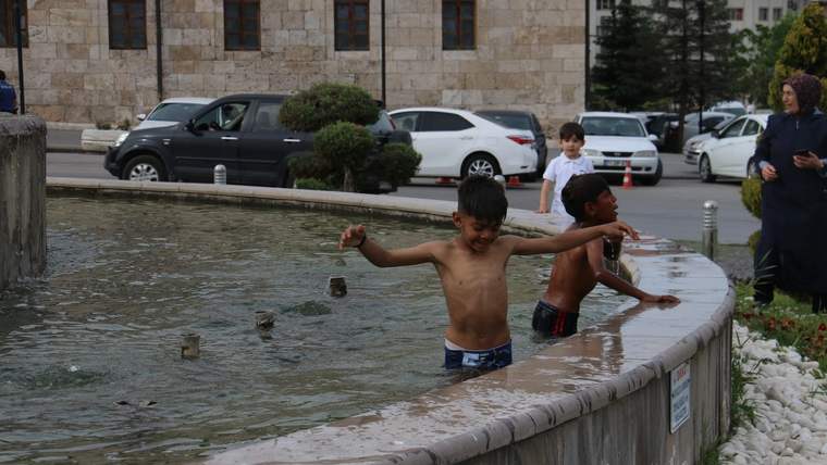 Süs havuzunda iki çocuk suyun içinde dururken, arkalarında araçlar ve taş kaplı bir bina görülüyor.