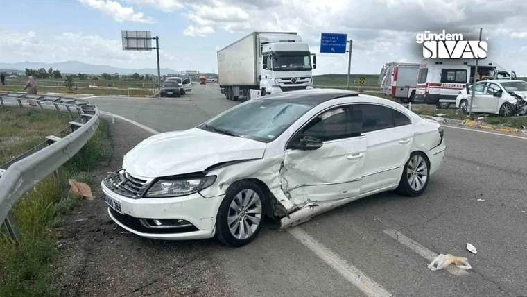 Hasarlı beyaz otomobil yol ayrımında duruyor; arka planda kamyon, ambulans ve başka araçlar görülüyor.