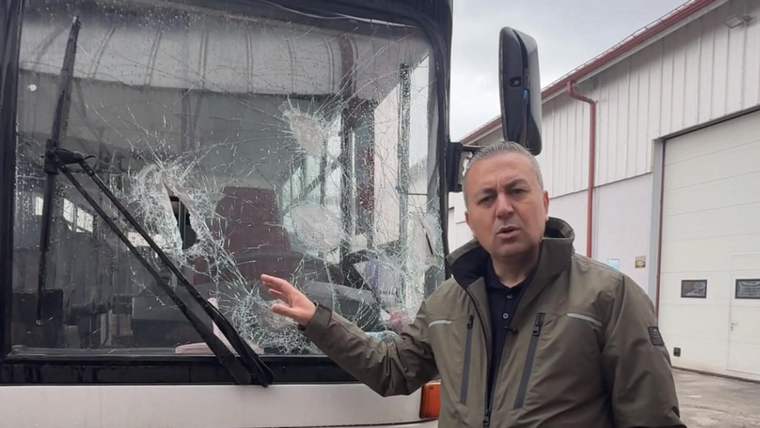 Bir kişi, camı kırılmış bir otobüsün önünde duruyor ve camı işaret ediyor.