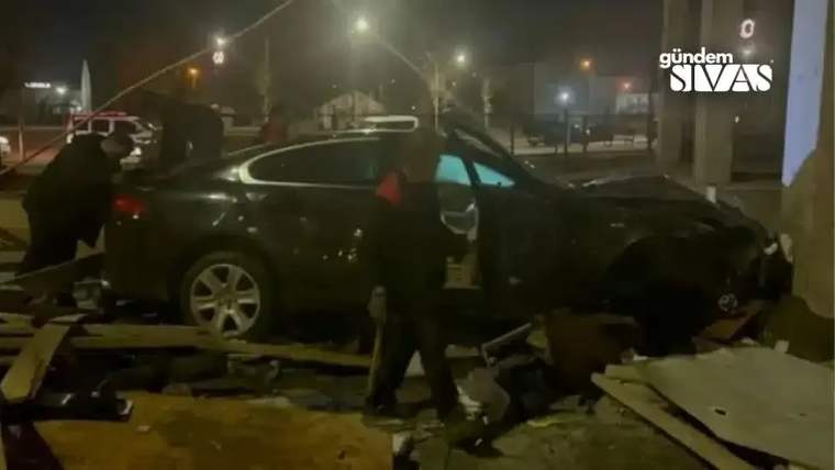 Gece, ön kısmı hasar gören siyah otomobilin çevresinde iki kişi ve beton parçaları görülüyor.