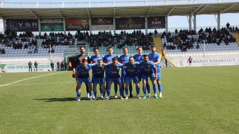 Futbol sahasında mavi formalar giyen 11 oyuncu, tribünlerin önünde takım fotoğrafı için poz veriyor.