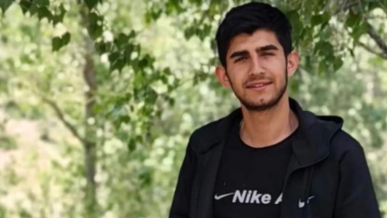 Yeşil ağaçların önünde duran genç bir adam, siyah tişört giymekte.