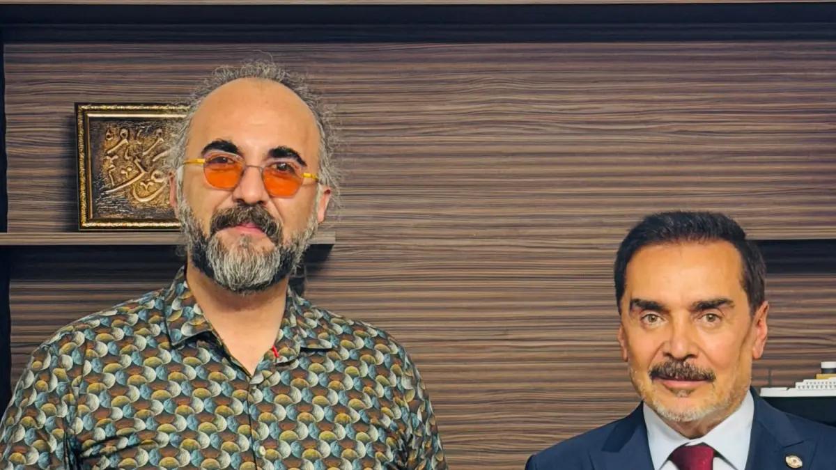 İki kişi, bir odada yan yana duruyor; arka planda ahşap bir raf ve bir tablo var.