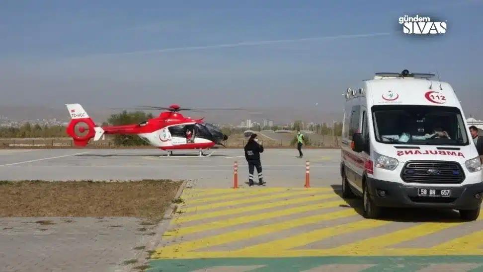 Kırmızı-beyaz bir ambulans ve helikopter, açık alanda duruyor; sağlık personeli helikoptere doğru yürüyor.