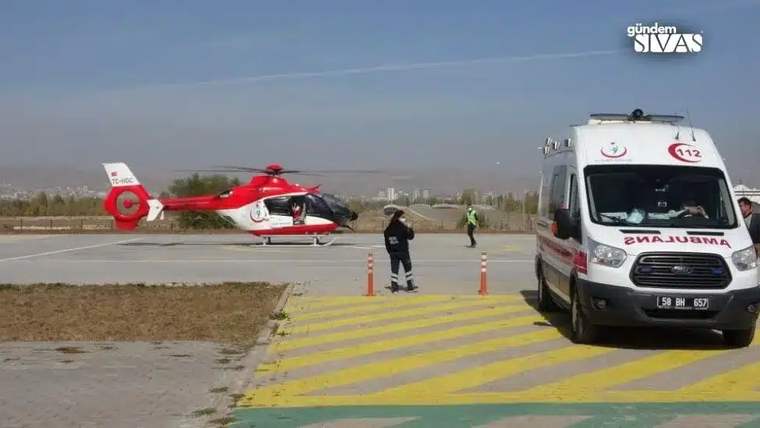 Kırmızı-beyaz bir ambulans ve helikopter, açık alanda duruyor; sağlık personeli helikoptere doğru yürüyor.