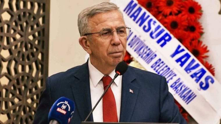 Mikrofon önünde konuşan gözlüklü bir kişi, takım elbise ve kırmızı kravat takıyor; arkasında çiçekler ve yazılı pankart var.