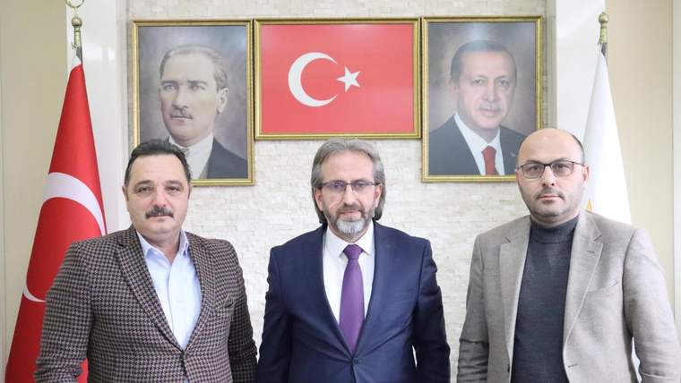 Üç erkek, Türk bayrakları ve iki portre arasında yan yana duruyor; ortadaki kişi takım elbise giyiyor.