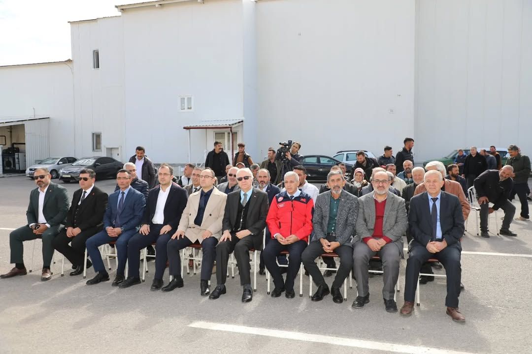 Beyaz bir binanın önünde, sandalyelerde oturan takım elbiseli kişiler ve arkalarında duran kalabalık görülüyor.