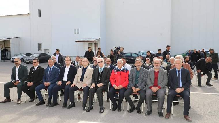 Beyaz bir binanın önünde, sandalyelerde oturan takım elbiseli kişiler ve arkalarında duran kalabalık görülüyor.