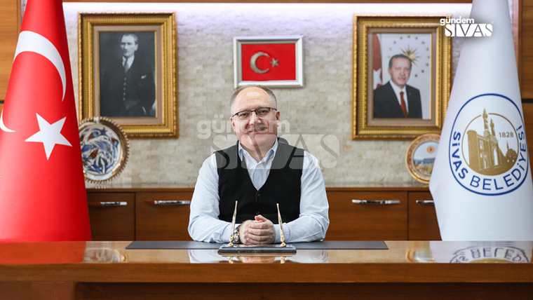 Gözlüklü bir kişi, Türk bayrağı ve belediye bayrağının yanında masada ellerini birleştirmiş oturuyor.