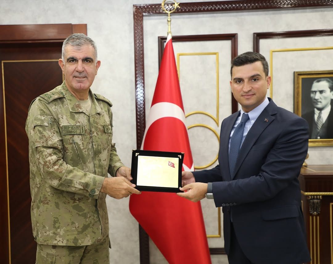 Askerî üniformalı bir kişi ile takım elbiseli bir kişi, Türk bayrağı önünde plaket tutuyor.