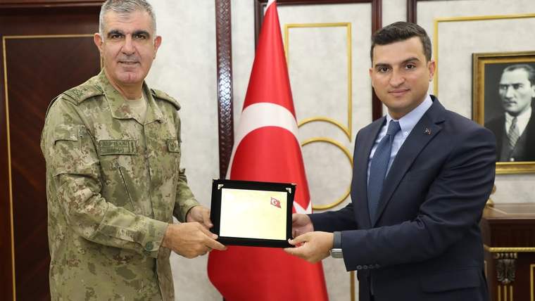 Askerî üniformalı bir kişi ile takım elbiseli bir kişi, Türk bayrağı önünde plaket tutuyor.