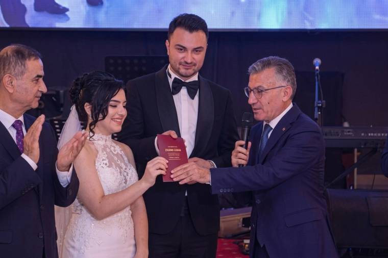 Bir salonda gelin ve damat kırmızı kitapçığı tutarken mikrofonlu bir erkek konuşuyor, yanlarında biri alkışlıyor.