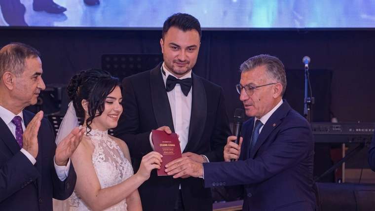 Bir salonda gelin ve damat kırmızı kitapçığı tutarken mikrofonlu bir erkek konuşuyor, yanlarında biri alkışlıyor.