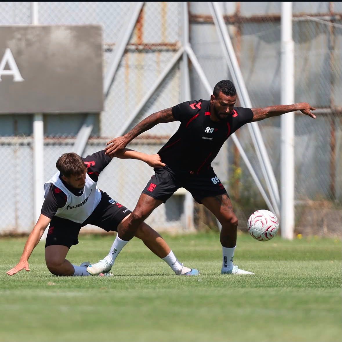 Çim sahada siyah formalı futbolcu topa uzanırken beyaz yelekli rakibi yere düşüyor.