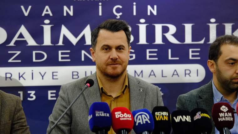 Mikrofonlar önünde konuşan bir kişi, arkasında “Van için” yazılı mor bir pankartla görülüyor.