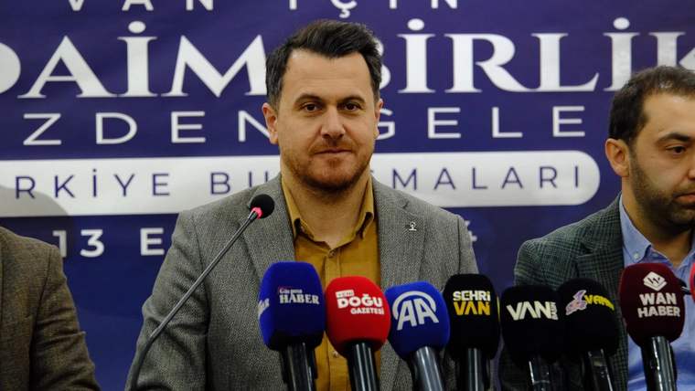 Kürsüde konuşan bir kişi, önünde çok sayıda mikrofon ve arkasında yazılı bir pankartla görülüyor.
