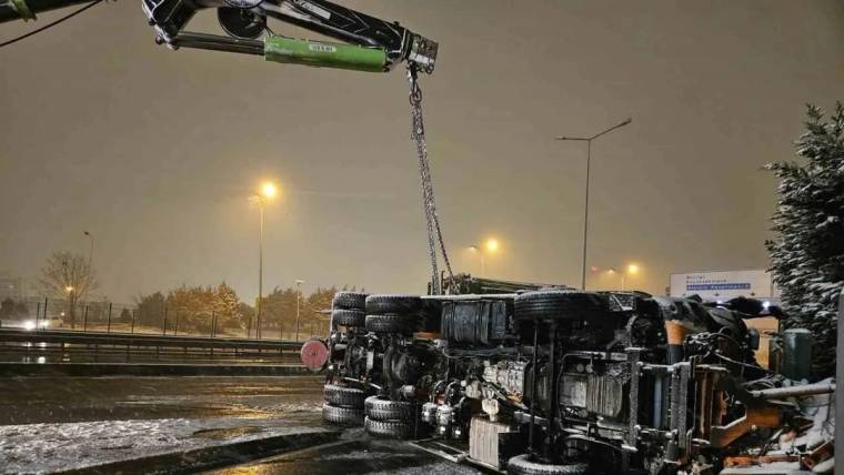 Gece karla kaplı yolda yan yatmış bir kamyon, vinçle kaldırılırken çevresinde ekipmanlar görülüyor.