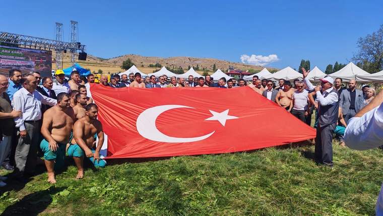 Çimenlik alanda kalabalık bir grup, önde güreşçilerle birlikte büyük Türk bayrağı tutuyor.