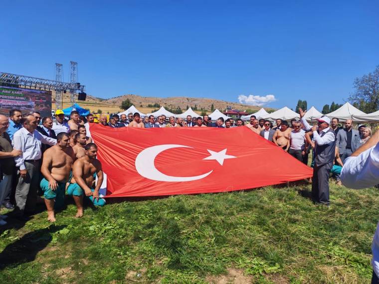 Çimenlik alanda kalabalık bir grup, önde güreşçilerle birlikte büyük Türk bayrağı tutuyor.