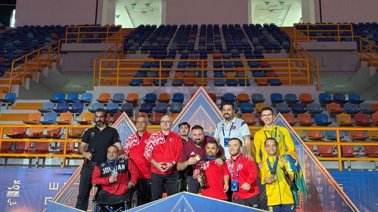 Spor salonunda, madalyalı sporcular ve görevliler, üzerinde World Para Powerlifting yazan platformda duruyor.