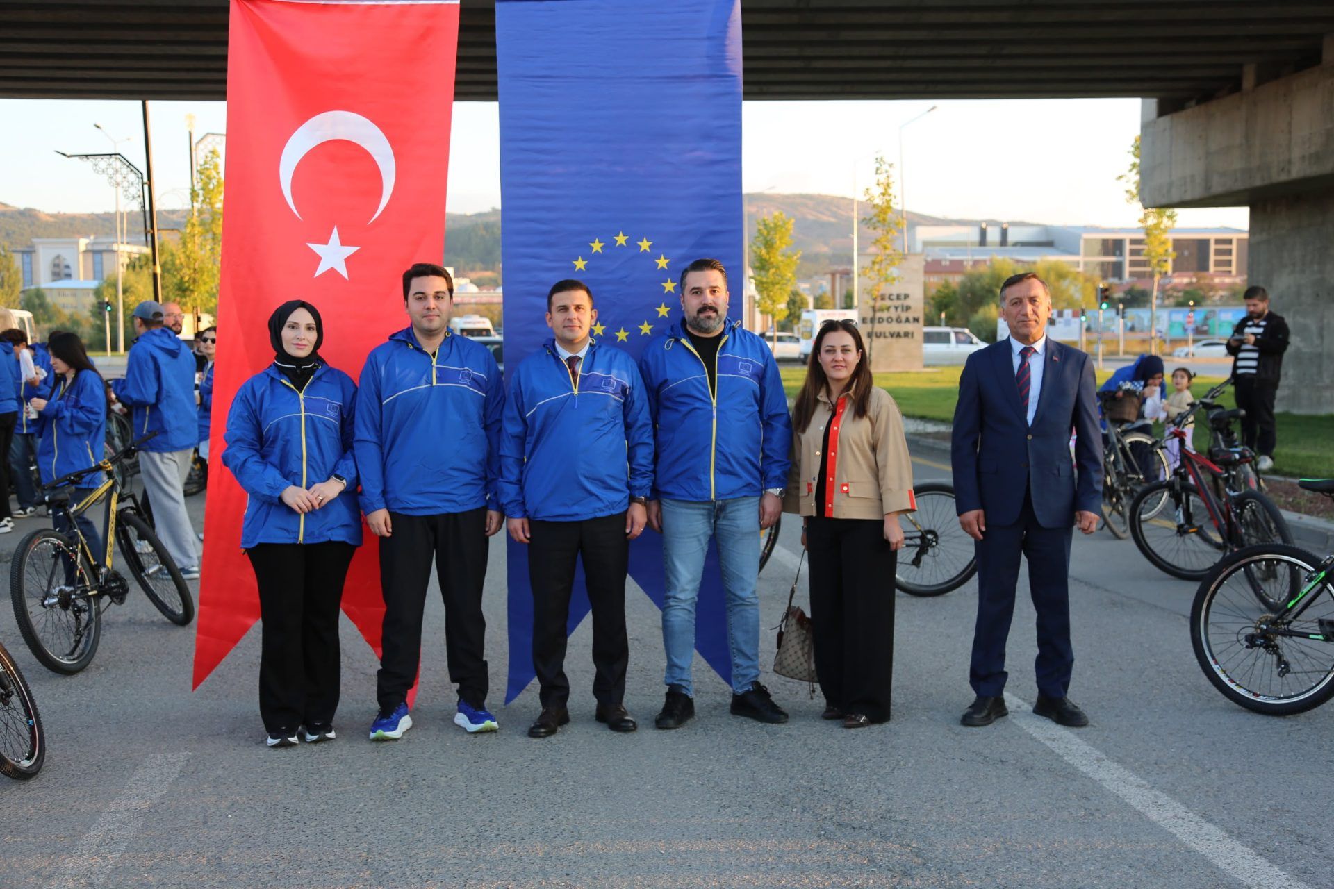 Türk ve Avrupa Birliği bayraklarının önünde, bisikletlerin arasında duran altı kişi birlikte poz veriyor.