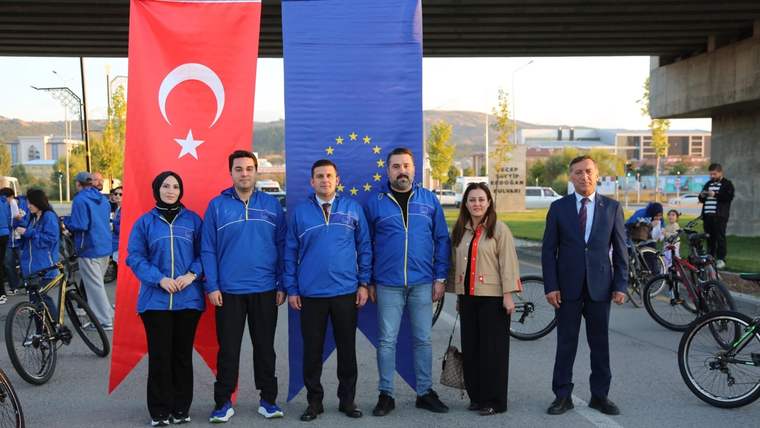 Türk ve Avrupa Birliği bayraklarının önünde, bisikletlerin arasında duran altı kişi birlikte poz veriyor.
