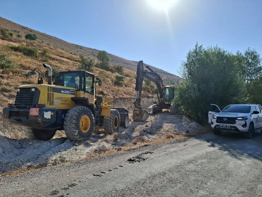 Yolda çalışan bir ekskavatör ve bir iş makinesi, yanlarında bir otomobil ile birlikte görüntüleniyor.