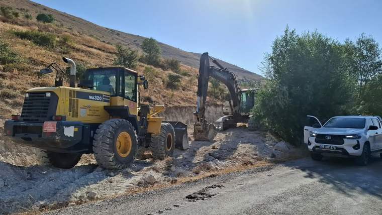 Yolda çalışan bir ekskavatör ve bir iş makinesi, yanlarında bir otomobil ile birlikte görüntüleniyor.