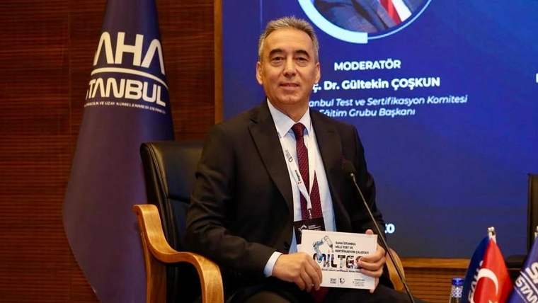 Takım elbiseli bir kişi, konferans salonunda elinde belgeyle oturuyor; yanında Türk bayrağı ve kürsü mikrofonu bulunuyor.