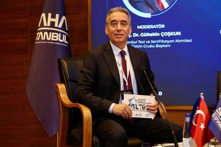 Takım elbiseli bir kişi, konferans salonunda elinde belgeyle oturuyor; yanında Türk bayrağı ve kürsü mikrofonu bulunuyor.