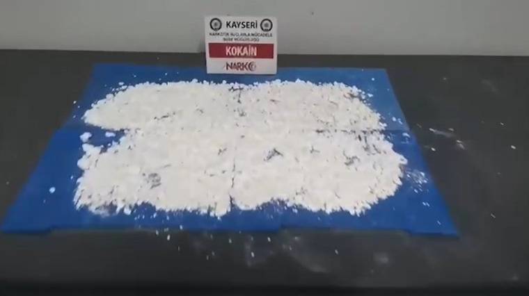 Mavi bir örtü üzerinde serili beyaz toz, arka planda 'KOKAİN' yazılı bir tabela var.