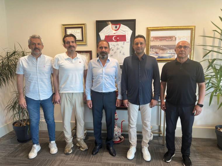 Duvarında çerçeveli belgeler ve forma bulunan odada beş erkek yan yana ayakta duruyor.