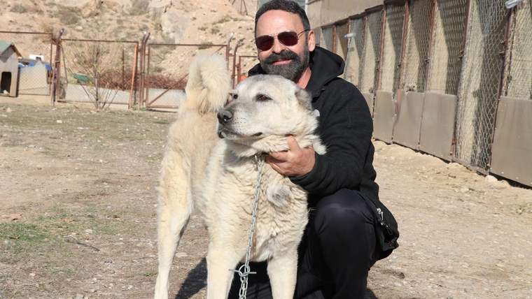 Kısa bir zincire bağlı bir köpeği seven bir adam, arka planda kafesler görünmekte.