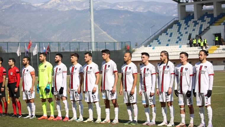 Futbol sahasında beyaz formalı oyuncular ve kırmızı formalı hakemler, tribünler önünde yan yana duruyor.
