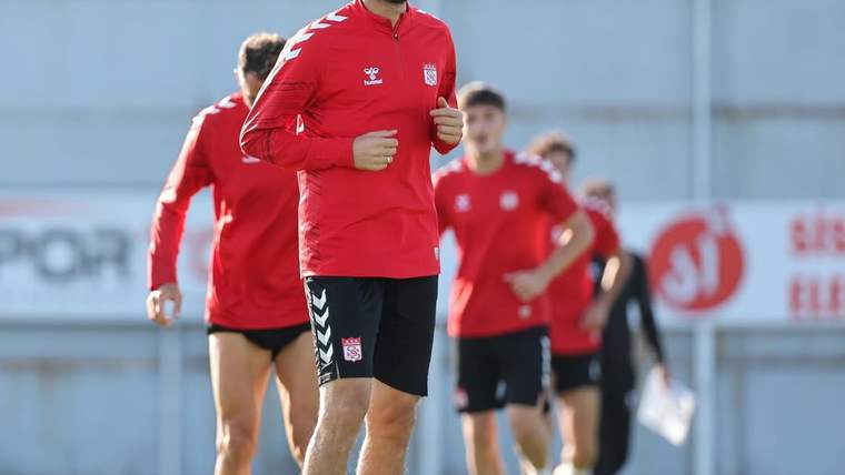 Kırmızı antrenman üstleri ve siyah şortlar giyen futbolcular, sahada sıra halinde koşu yapıyor.