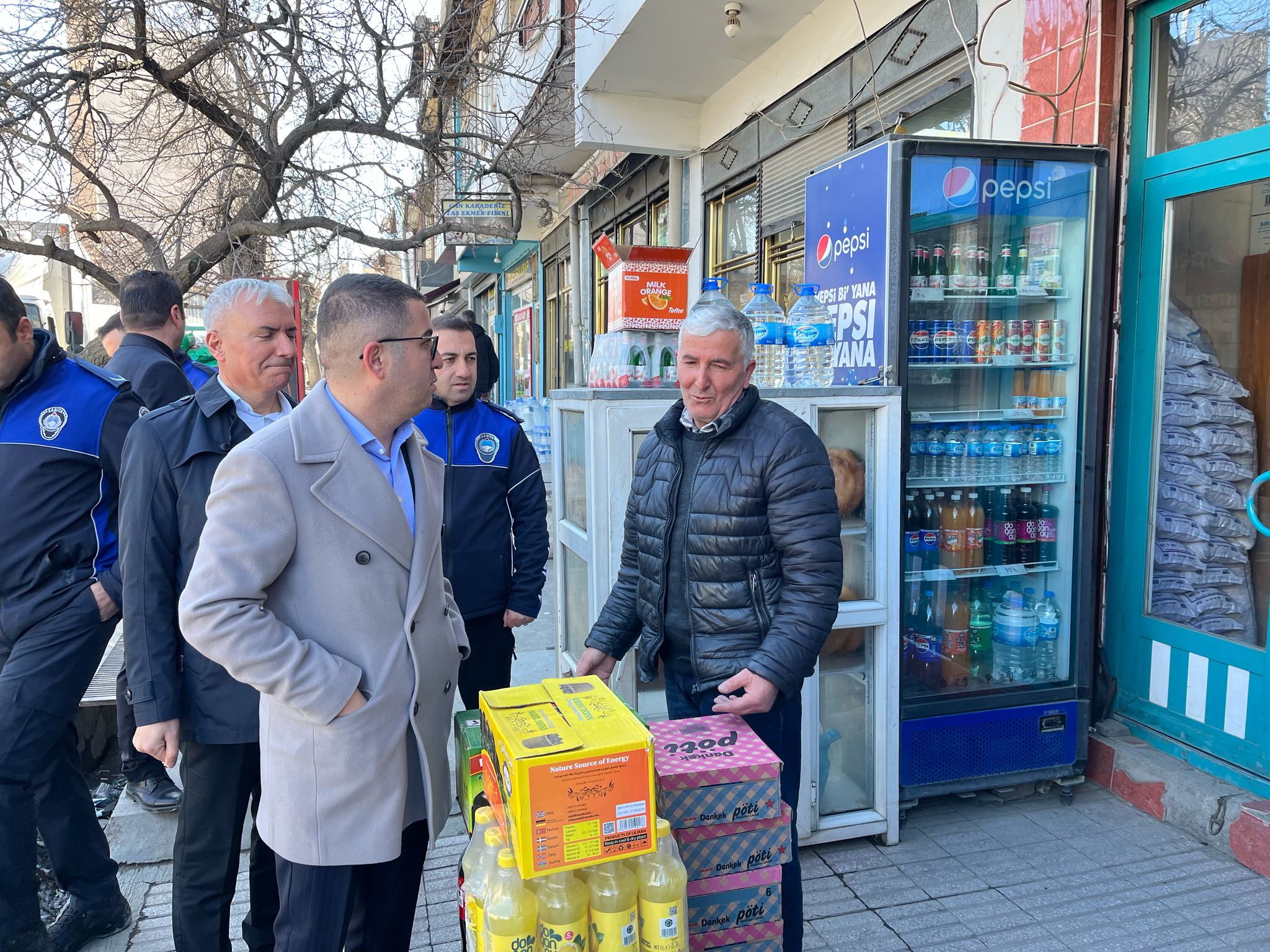 Bir dükkânın önünde montlu ve üniformalı kişiler, içecek dolabının yanında duran kolilere bakıyor.