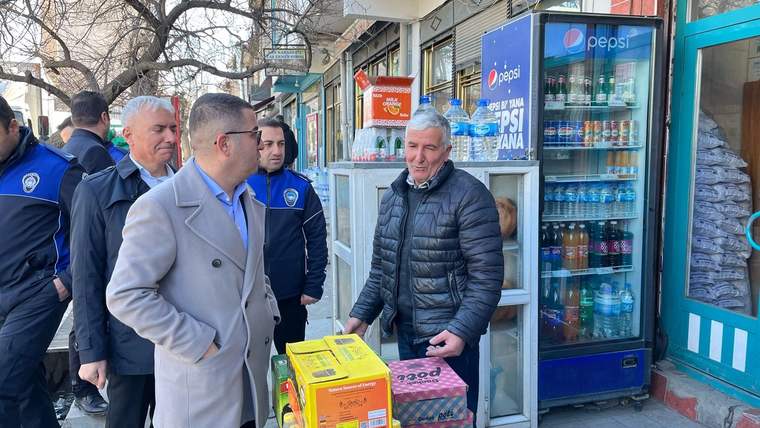 Bir dükkânın önünde montlu ve üniformalı kişiler, içecek dolabının yanında duran kolilere bakıyor.