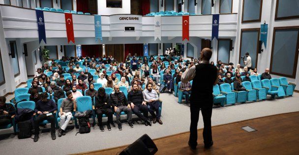 Bir kişi sahnede konuşurken, dinleyiciler mavi koltuklarda oturuyor.