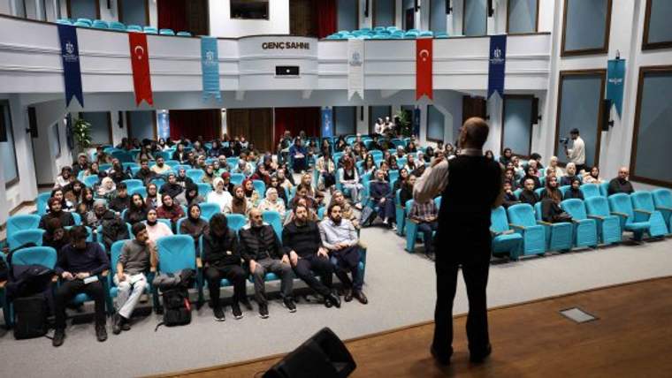 Bir kişi sahnede konuşurken, dinleyiciler mavi koltuklarda oturuyor.