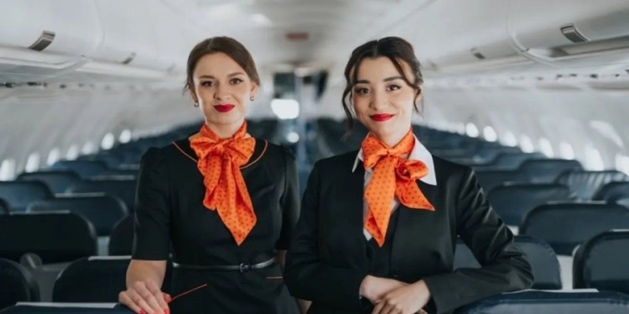 Uçak kabininde siyah üniformalı ve turuncu fularlı iki kadın yolcu koltuklarının önünde duruyor.