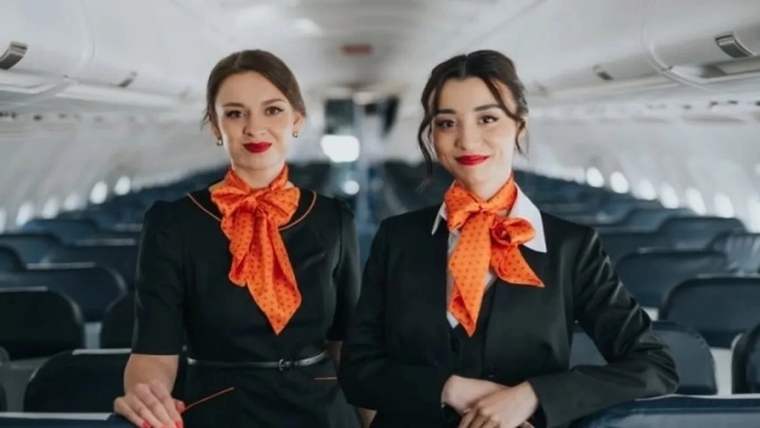 Uçak kabininde siyah üniformalı ve turuncu fularlı iki kadın yolcu koltuklarının önünde duruyor.