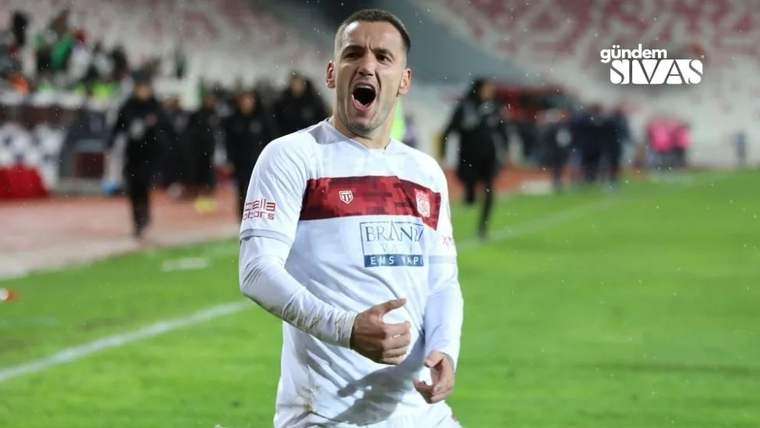 Yeşil saha kenarında beyaz-kırmızı formasıyla bir futbolcu, ağzı açık ve elleriyle sevinç işareti yapıyor.
