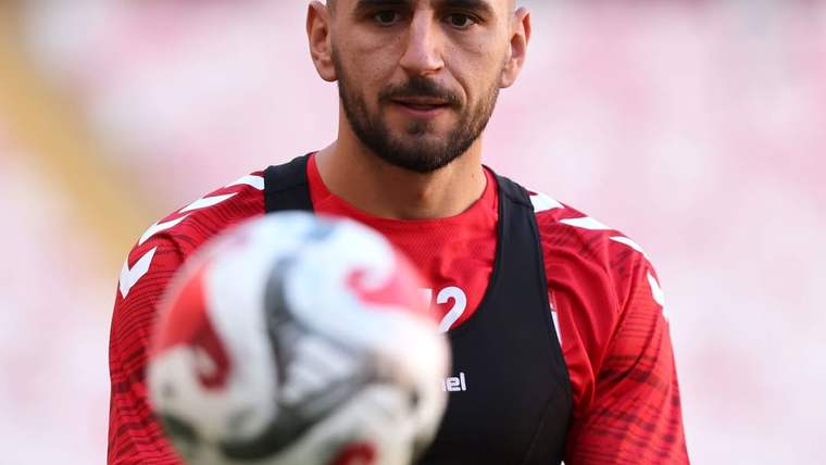 Kırmızı formalı ve siyah antrenman yelekli bir futbolcu, ön plandaki topun arkasında duruyor.